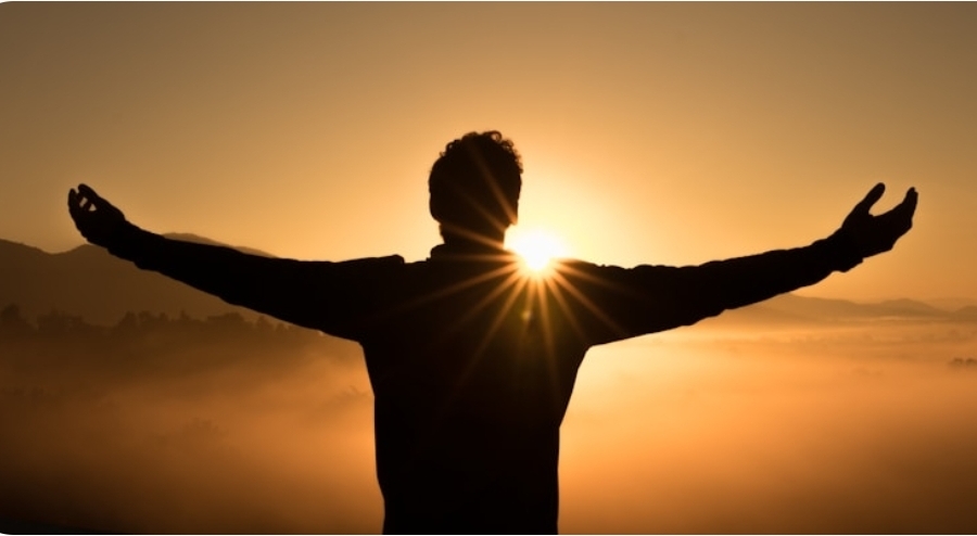 A person standing on a hilltop at sunrise with arms open wide, silhouetted against the light